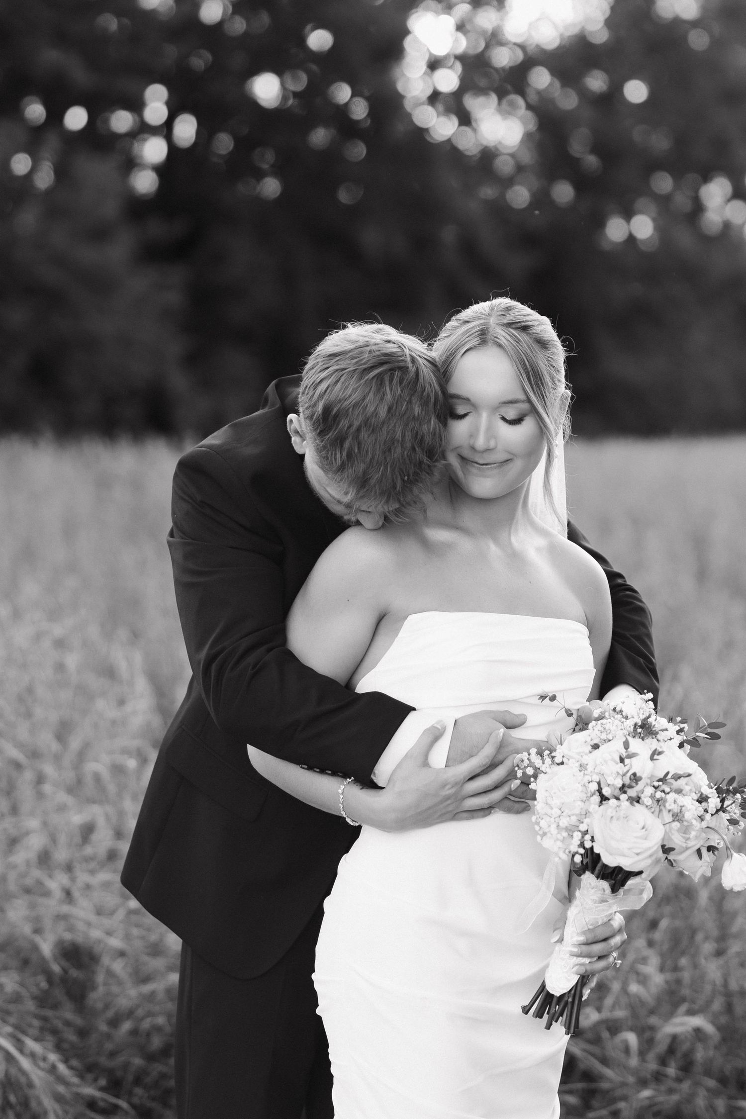Black and white intimate portrait of newlyweds embracing in field with bouquet.