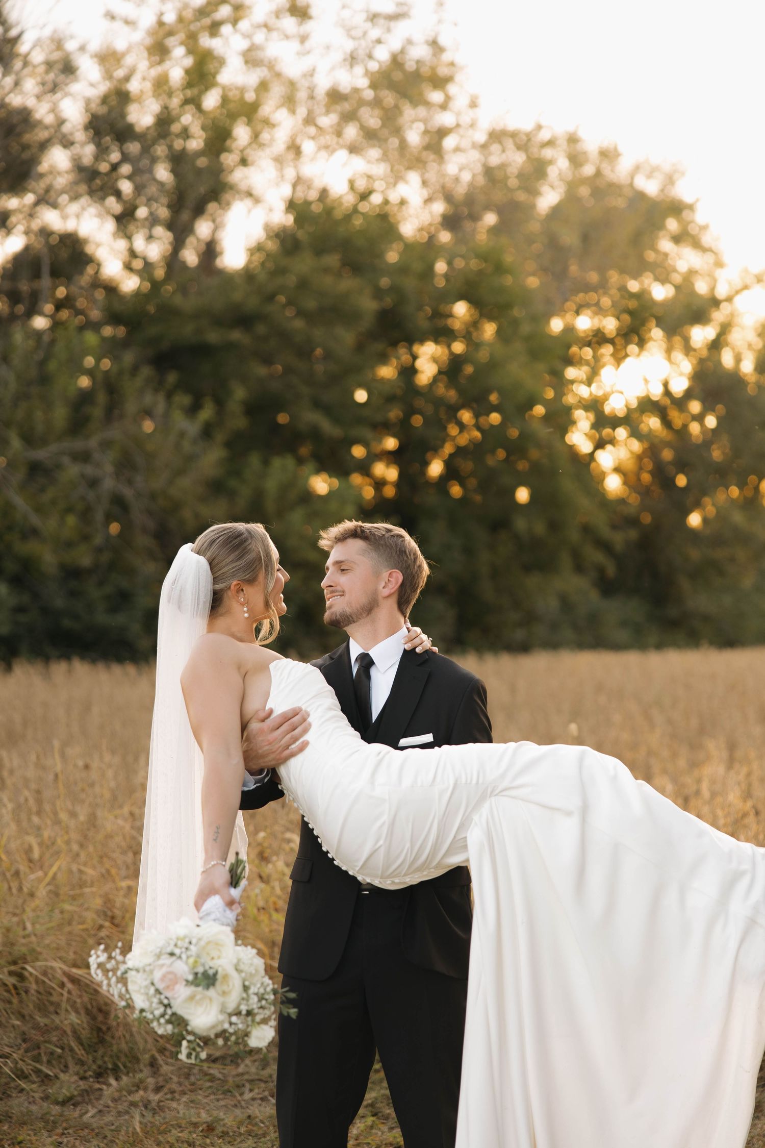 Groom carries bride in white dress during sunset portrait session in rustic field.