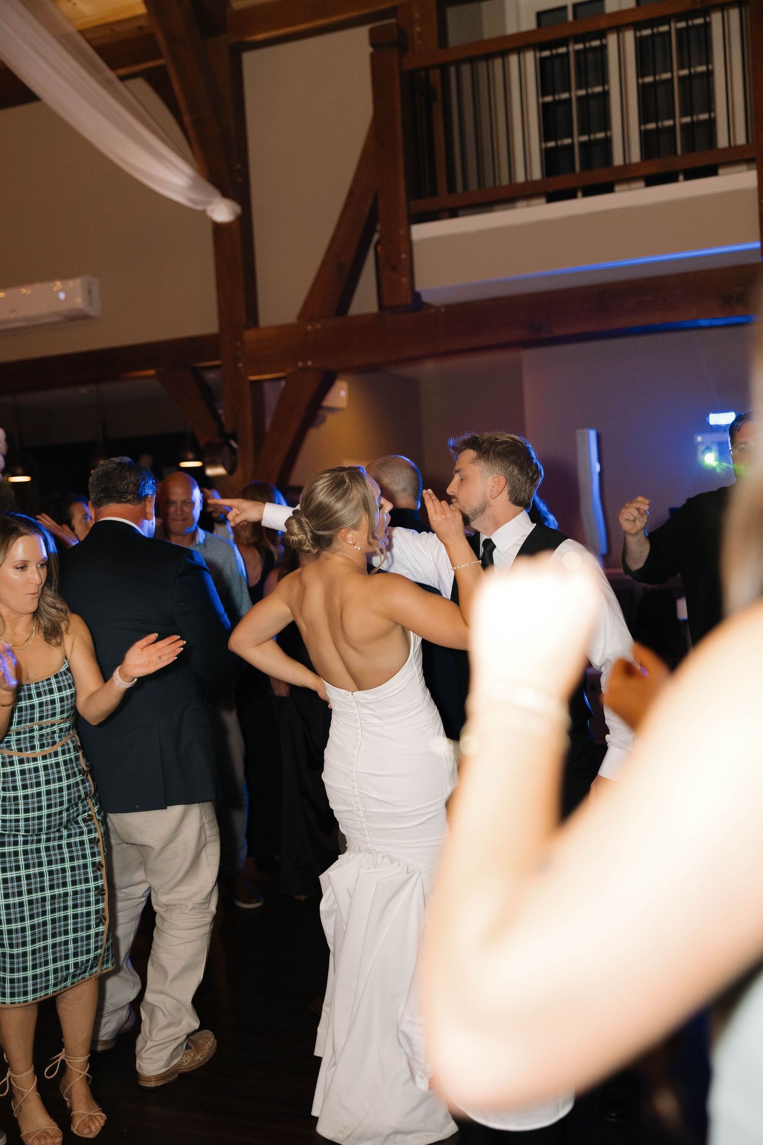 People dancing and having fun at a wedding reception with blue mood lighting and wooden beams visible.