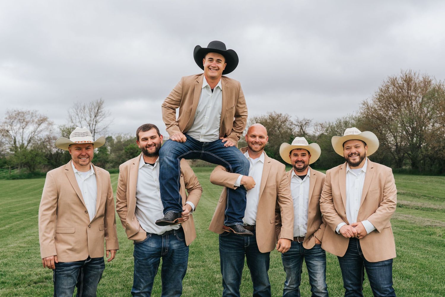 A group in matching beige blazers and cowboy hats poses together on a grassy field during a rustic outdoor celebration.