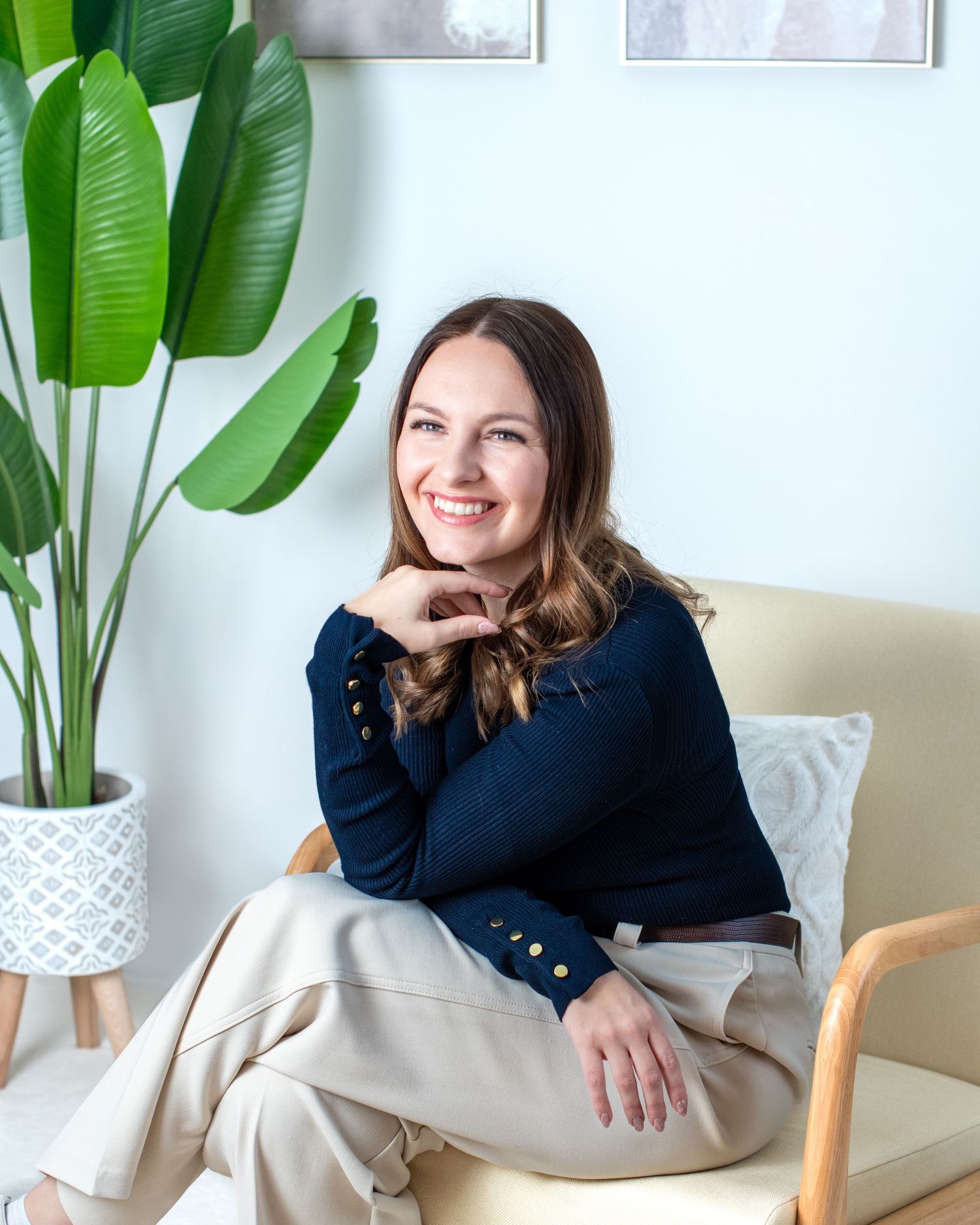 Woman in navy sweater sits comfortably in modern chair with large tropical plants and white planter in bright room.