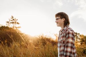 Person in plaid shirt stands in golden sunlit field during sunset while looking away from camera.