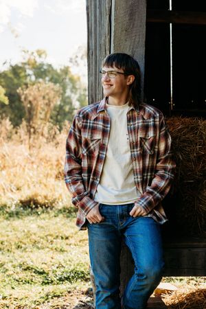 Person in plaid flannel shirt and jeans leans against barn door frame in golden sunlit rural setting.