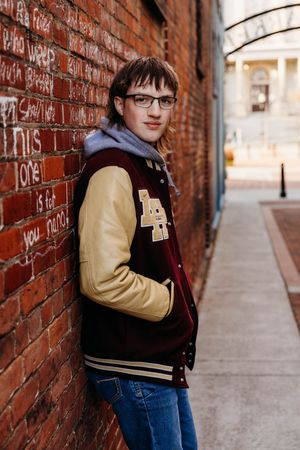 A person wearing a varsity jacket and glasses poses against a brick wall in an alley with graffiti.