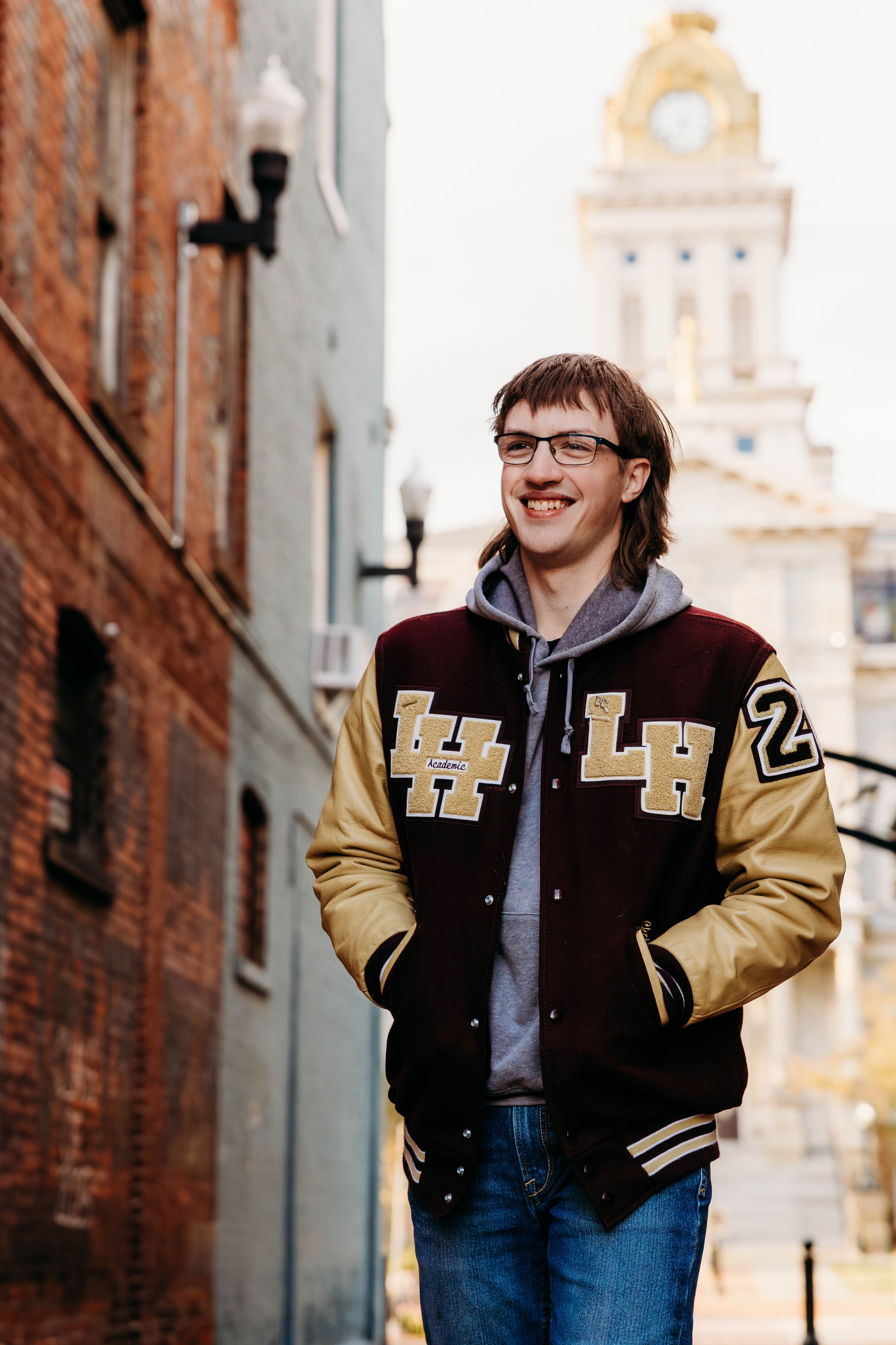A person in a varsity jacket and glasses poses against a brick wall backdrop in an urban alleyway.