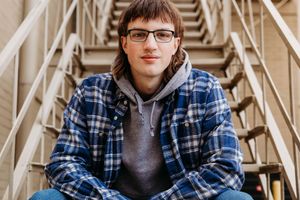 A person in a blue plaid shirt and glasses sits in front of a metallic industrial background.