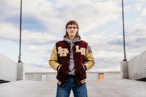 Person in a varsity jacket and jeans stands confidently against an urban backdrop with cloudy skies.