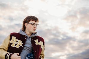 A person wearing a varsity letterman jacket with H patches stands against a cloudy sky background.