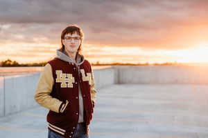 A person in a varsity jacket stands on a rooftop during a golden sunset with dramatic clouds in the sky.