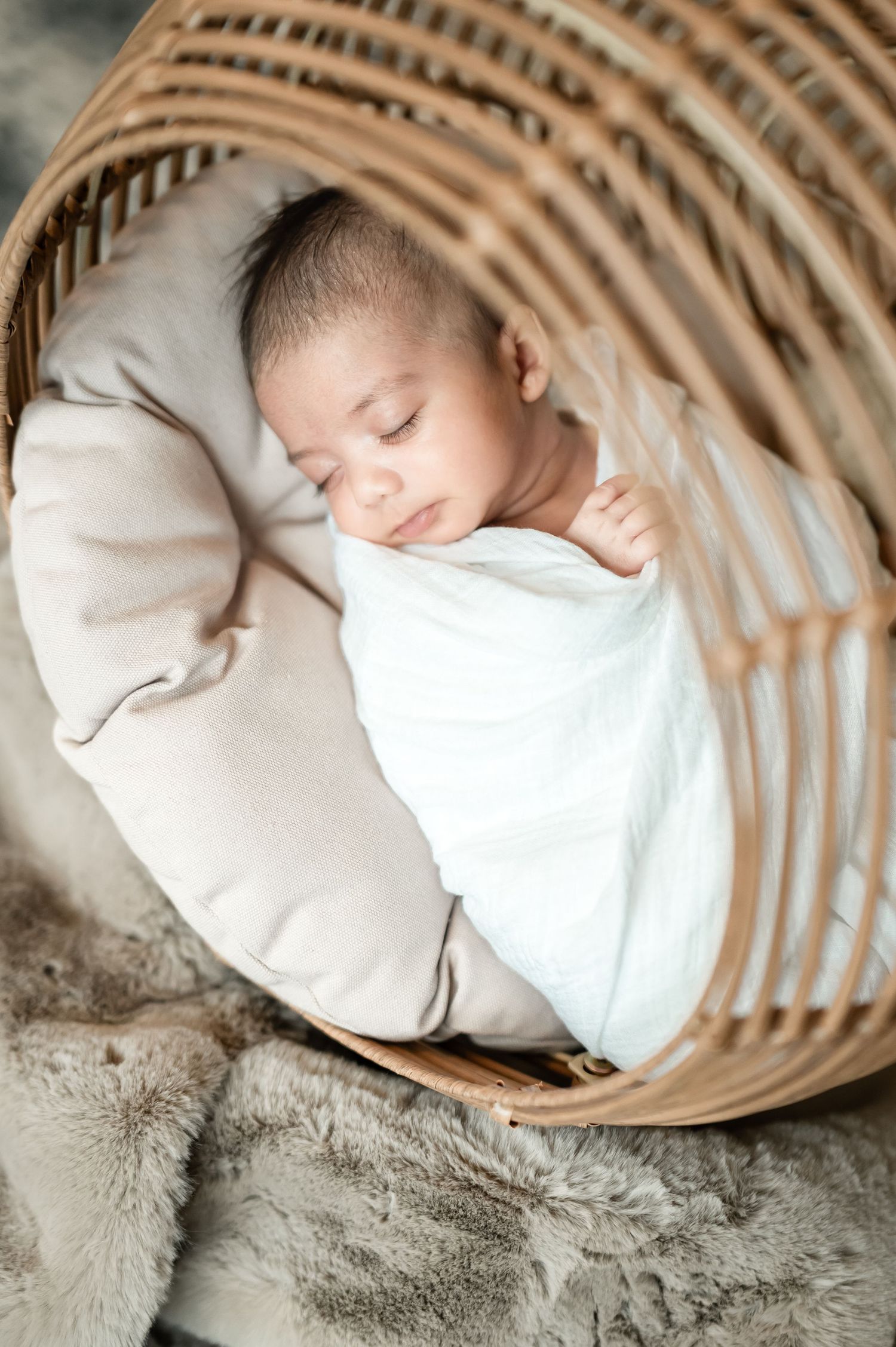 Sleeping newborn baby wrapped in white and beige blankets nestled in woven basket on soft gray fur backdrop.