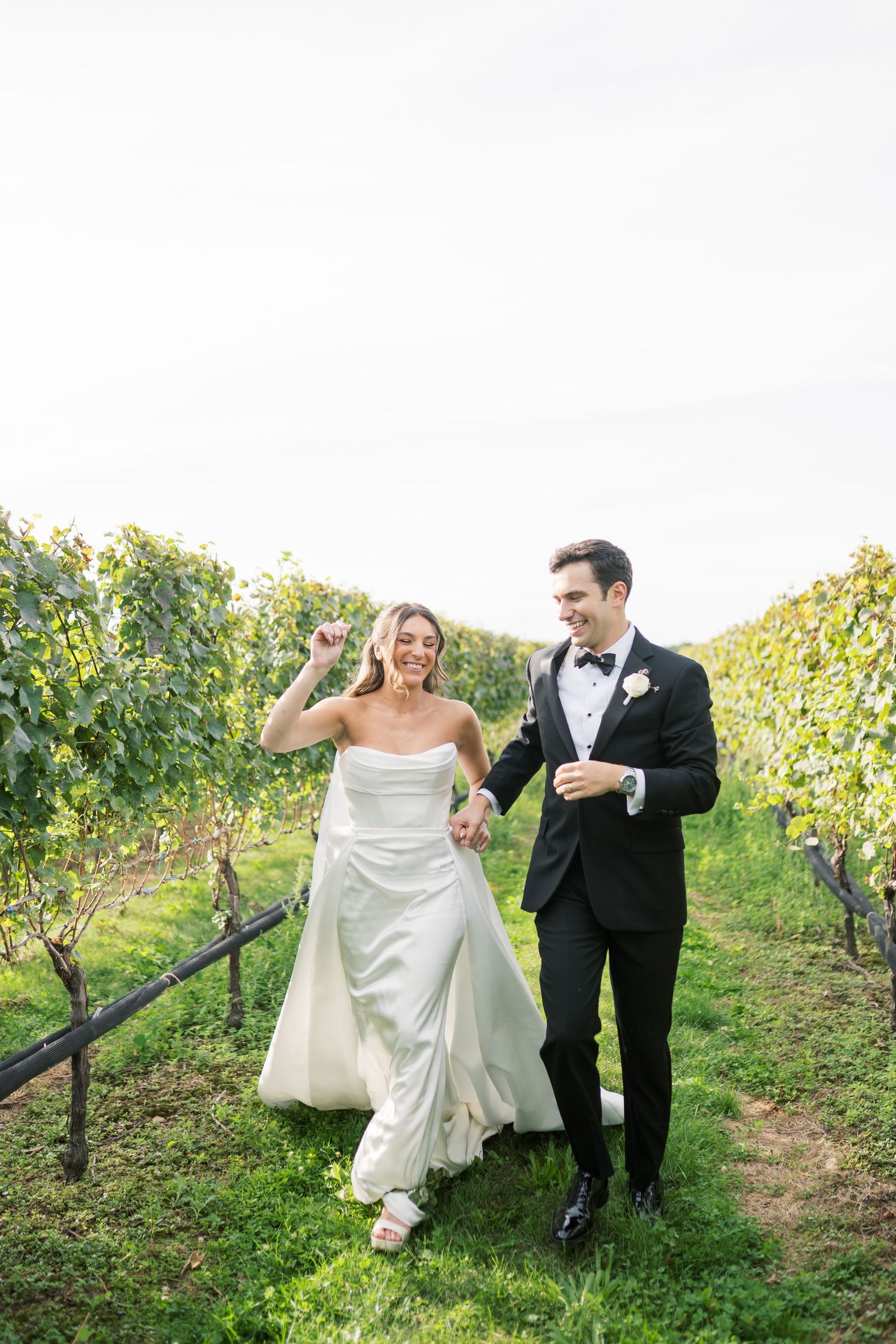 A couple walks together through scenic vineyard rows on their wedding day, with the bride in white and groom in black.