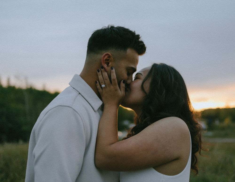 A romantic couple shares a tender kiss in a field at sunset while wearing white outfits.