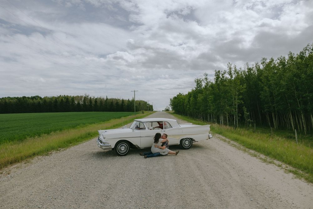 Classic car parked on a gravel road with green fields and trees in the background under cloudy skies.