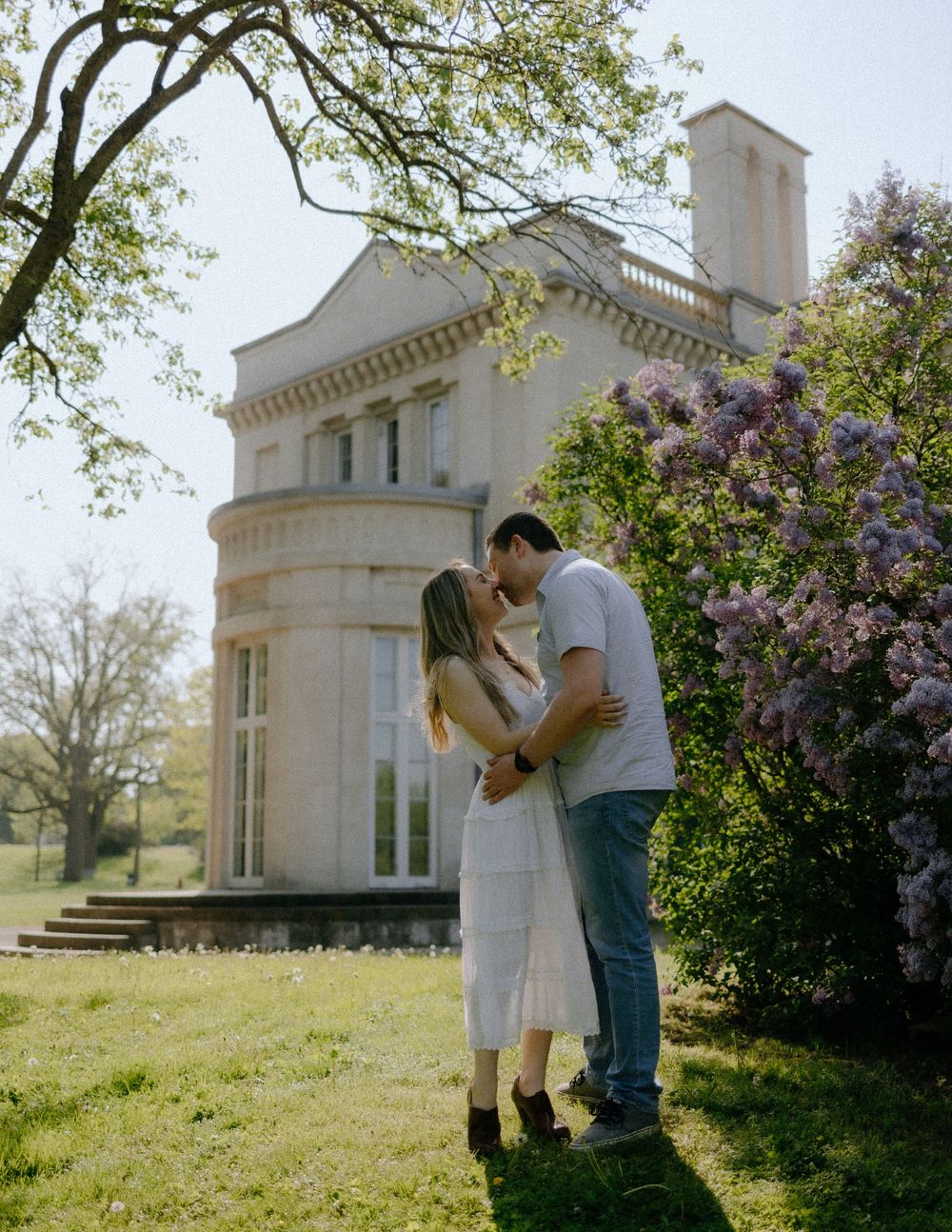 A romantic photo session takes place near a white classical building with blooming lilac trees in spring.