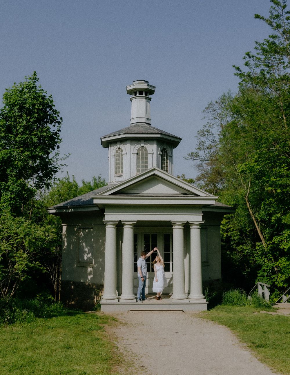 A white neoclassical pavilion with columns sits atop a grassy hill on a sunny spring day.