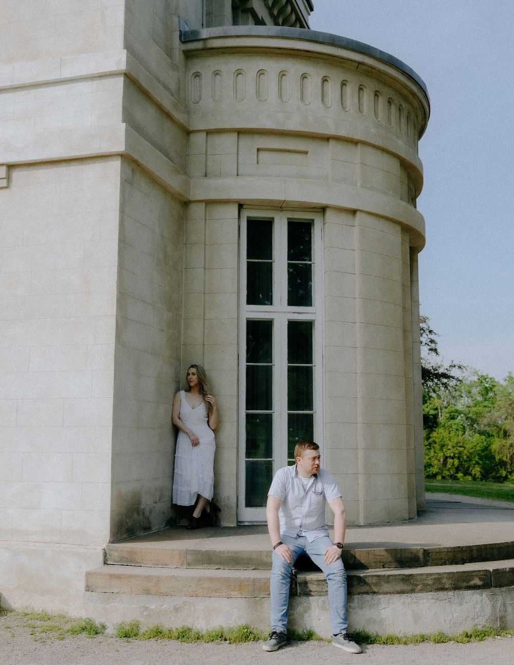 Two people pose by a curved white stone building with tall windows and architectural details.