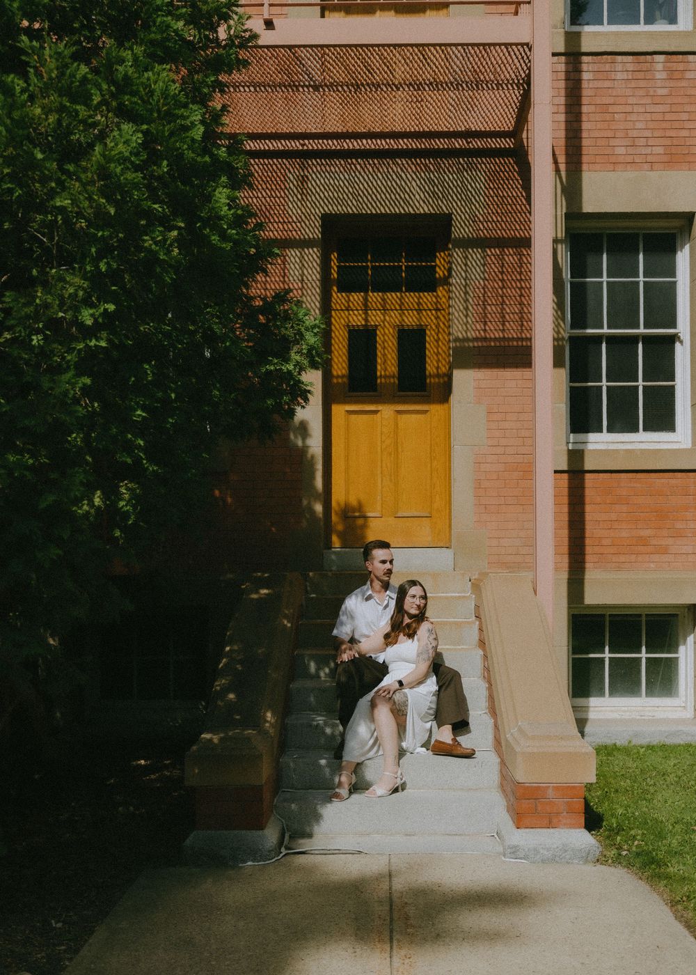 A couple sits together on concrete steps leading to a yellow door of a brick building on a sunny day.