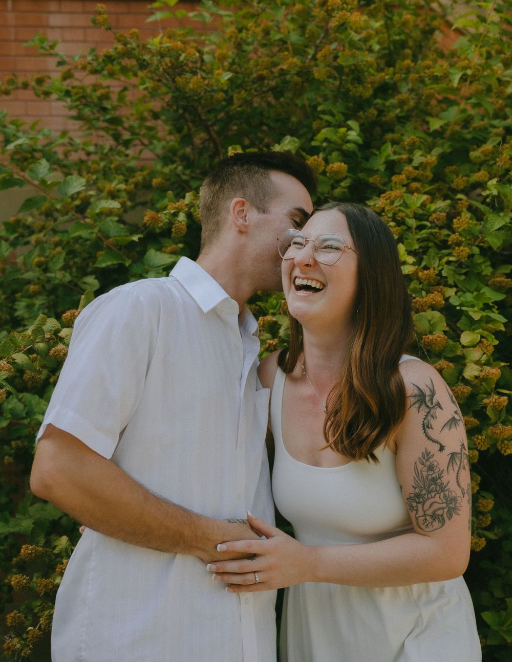 A couple shares intimate moments and laughter while posing together against a backdrop of green foliage.