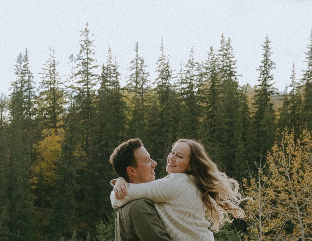 A couple shares a romantic moment in a forest with tall evergreen trees during golden hour.