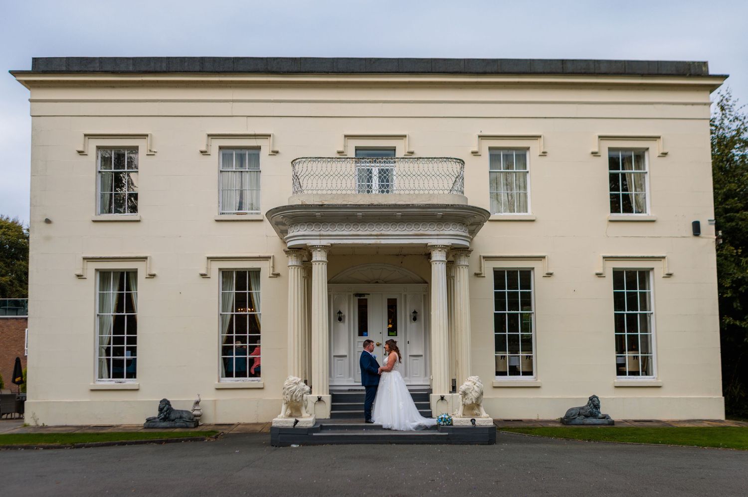 Bride and groom pose outside an elegant cream Georgian manor house with columns and balcony on their wedding day.