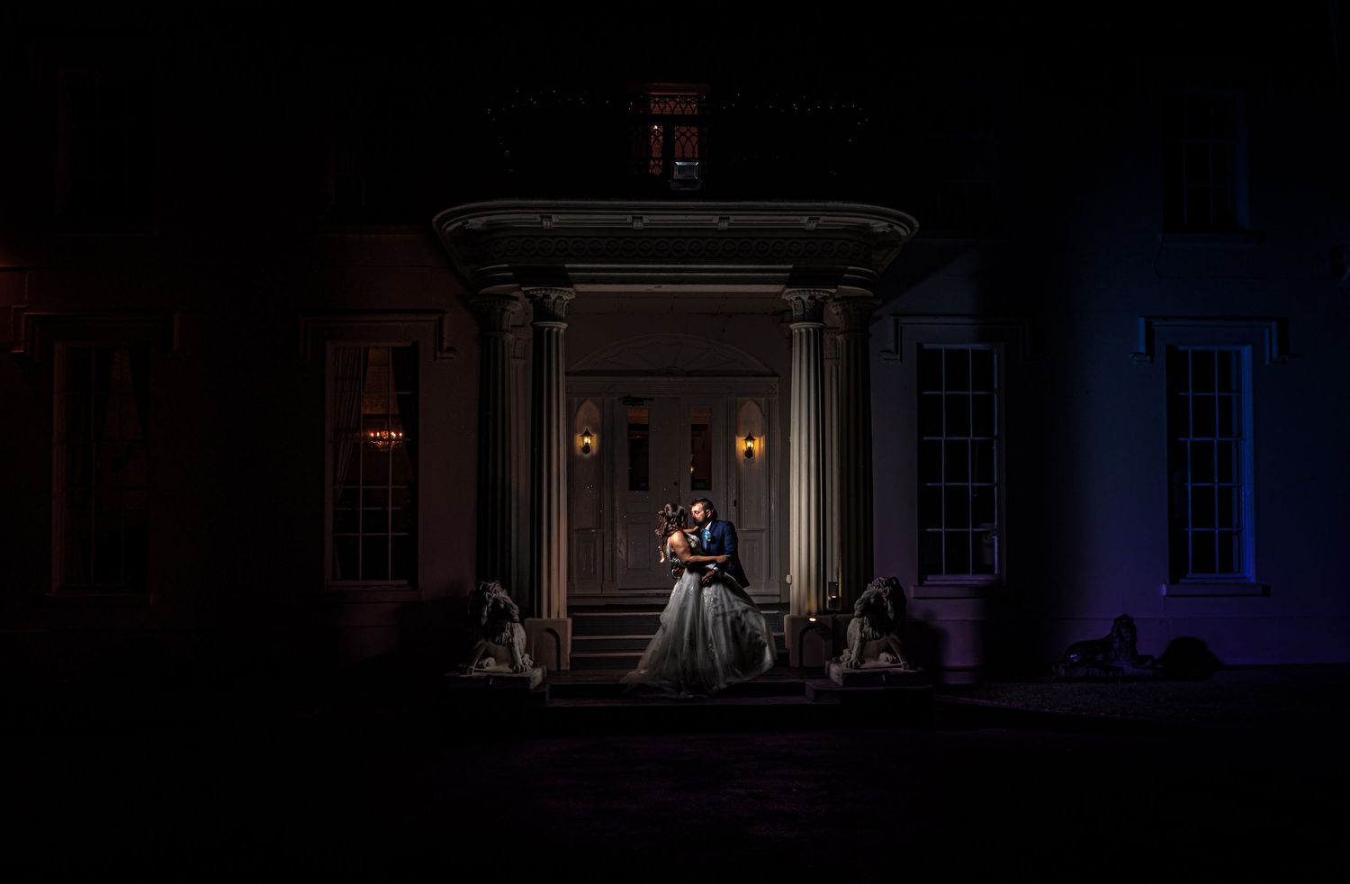 A dramatic nighttime wedding photo on a mansion's front porch illuminated by spotlights against a dark background.