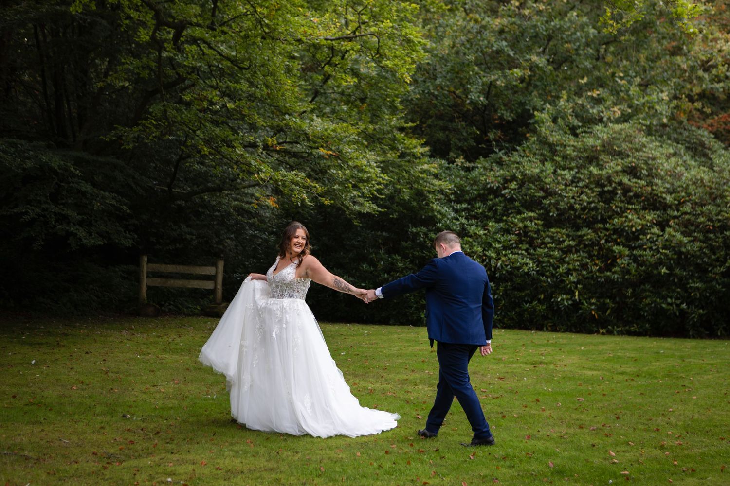 A couple dances together on a green lawn surrounded by lush forest trees during their outdoor wedding celebration.