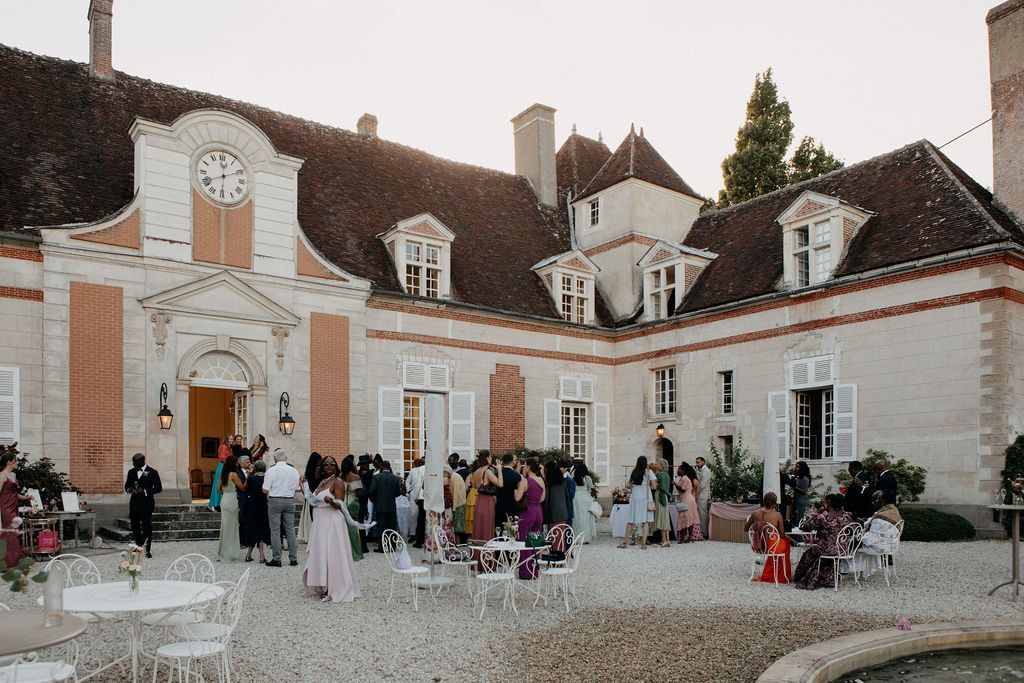 Elegant French chateau wedding reception with guests mingling in a courtyard featuring white furniture and a clock tower.