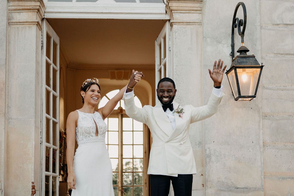 Newlyweds celebrating at chateau entrance raising hands in joy after wedding ceremony.