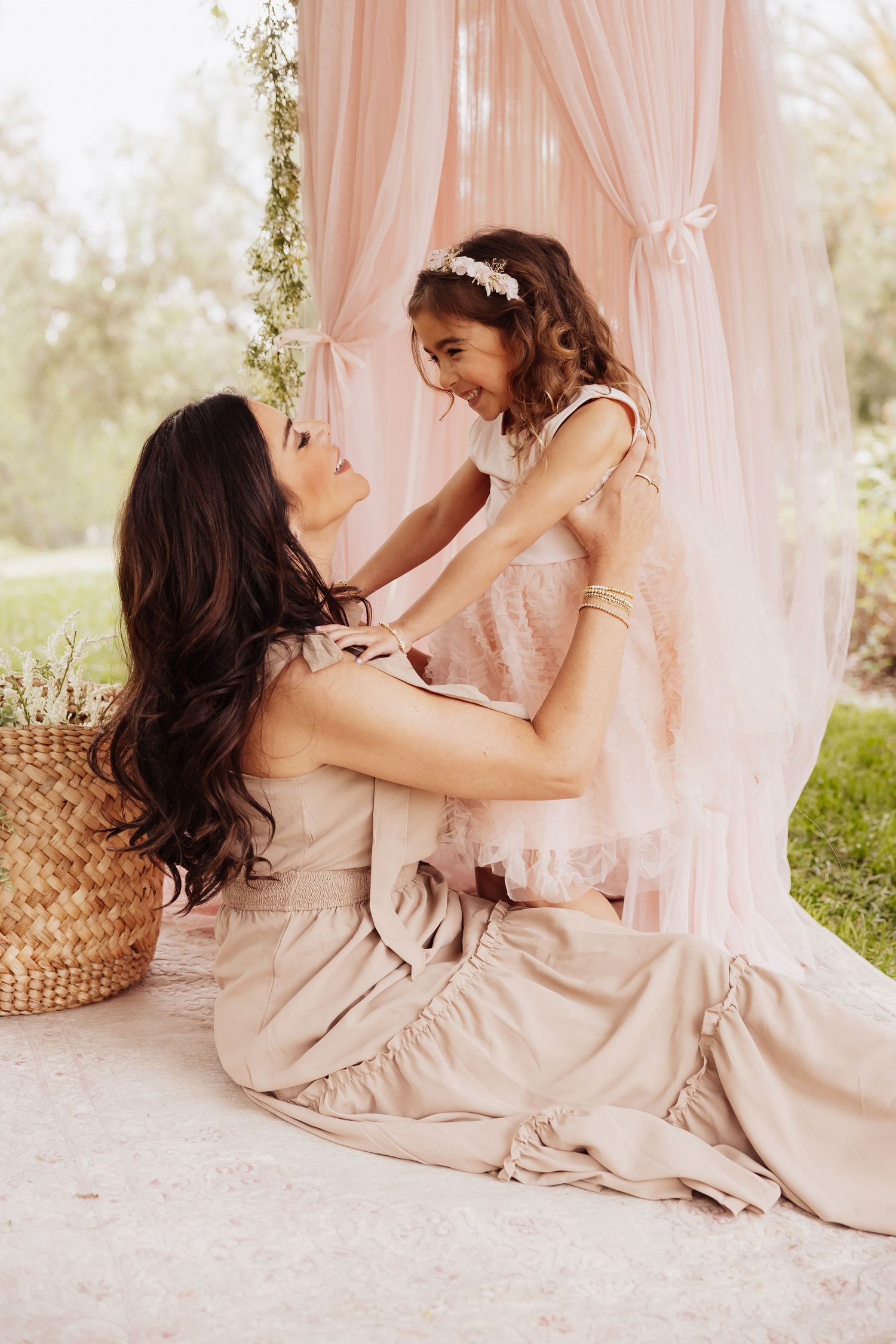 A tender moment captured between parent and child wearing blush-toned dresses against a soft pink draped backdrop outdoors.