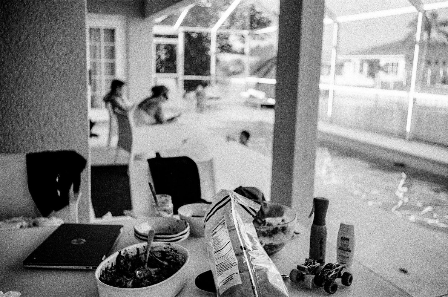 A black and white film photograph shows everyday objects on a table with figures silhouetted in the background near windows.