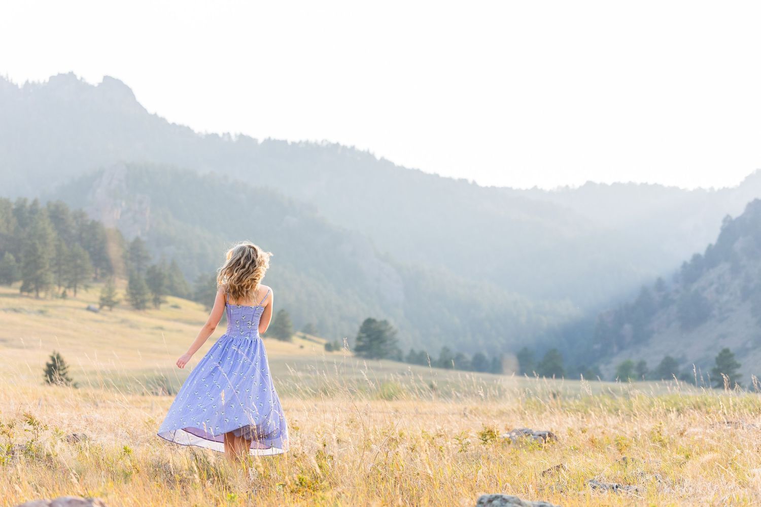 Woman in white dress stands outdoors with mountain landscape and blurred town in background.