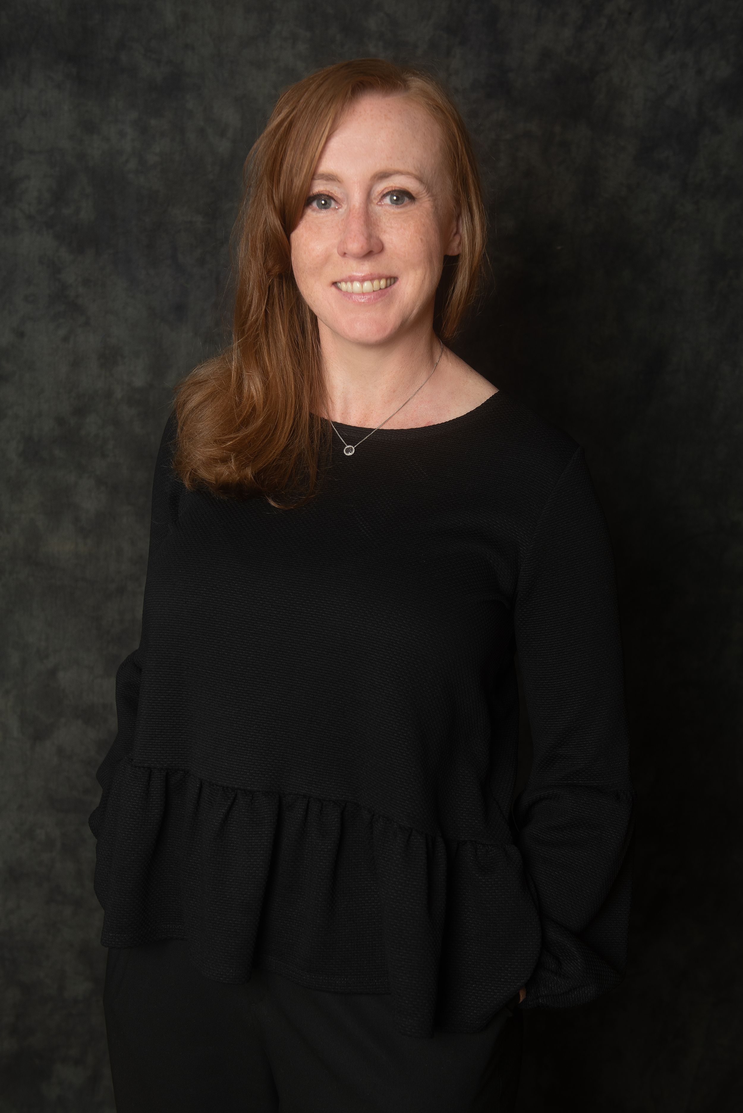 Professional headshot of someone wearing a black top against dark backdrop.