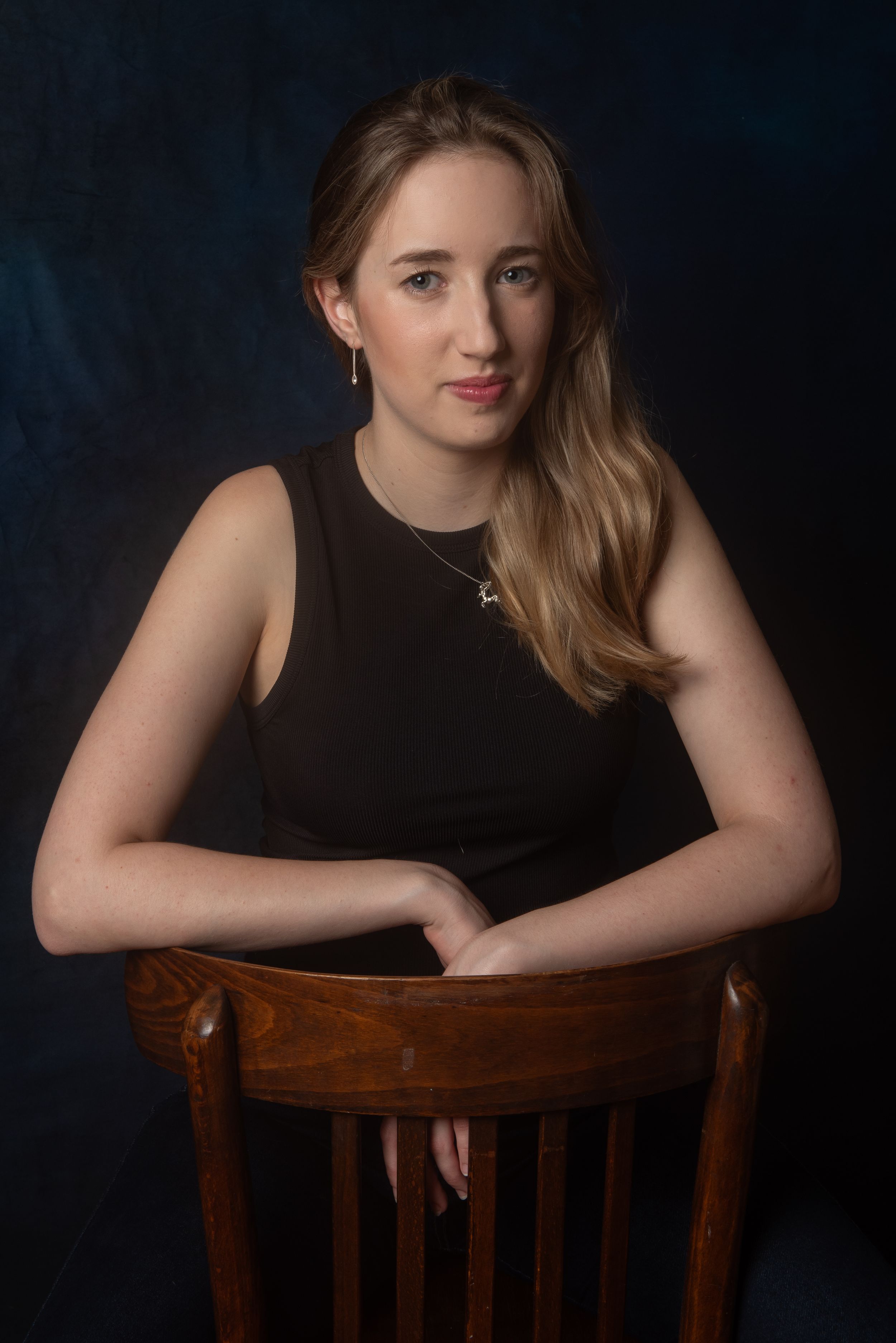 Person in black sleeveless top posed on wooden chair in studio setting.
