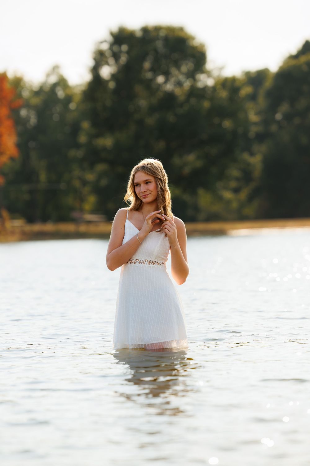 A person in a white dress stands in calm lake water during a golden hour photoshoot.