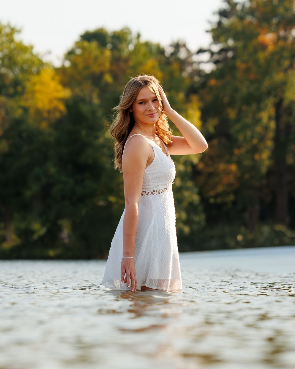 A woman in a white dress stands in shallow lake water during golden hour with trees in the background.