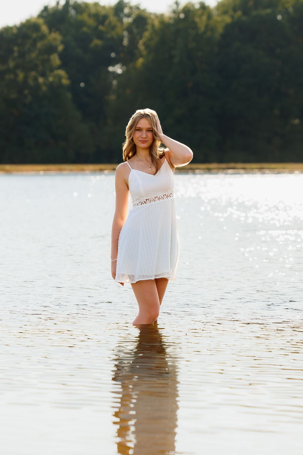 A person in a white top stands in shallow lake water during golden hour creating a serene reflection.