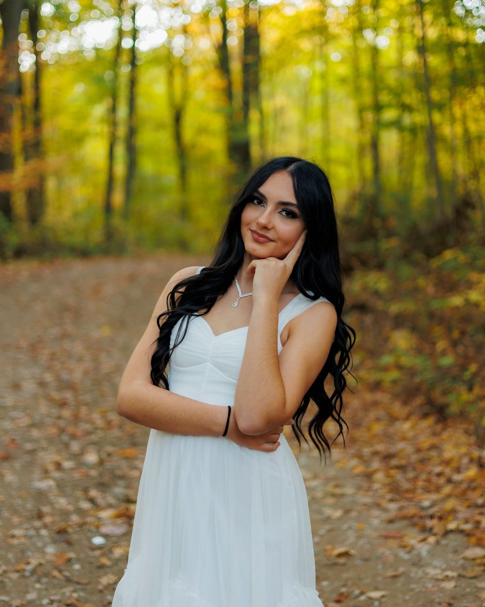 A figure in a flowing white dress poses gracefully among autumn leaves in a sunlit forest setting.