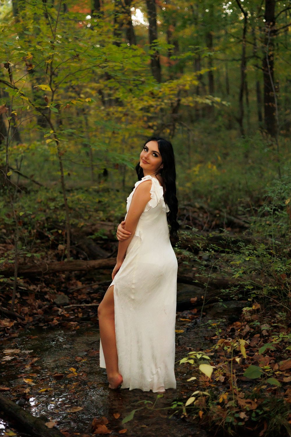 A figure in a flowing white dress poses gracefully among autumn trees and fallen leaves in a forest setting.