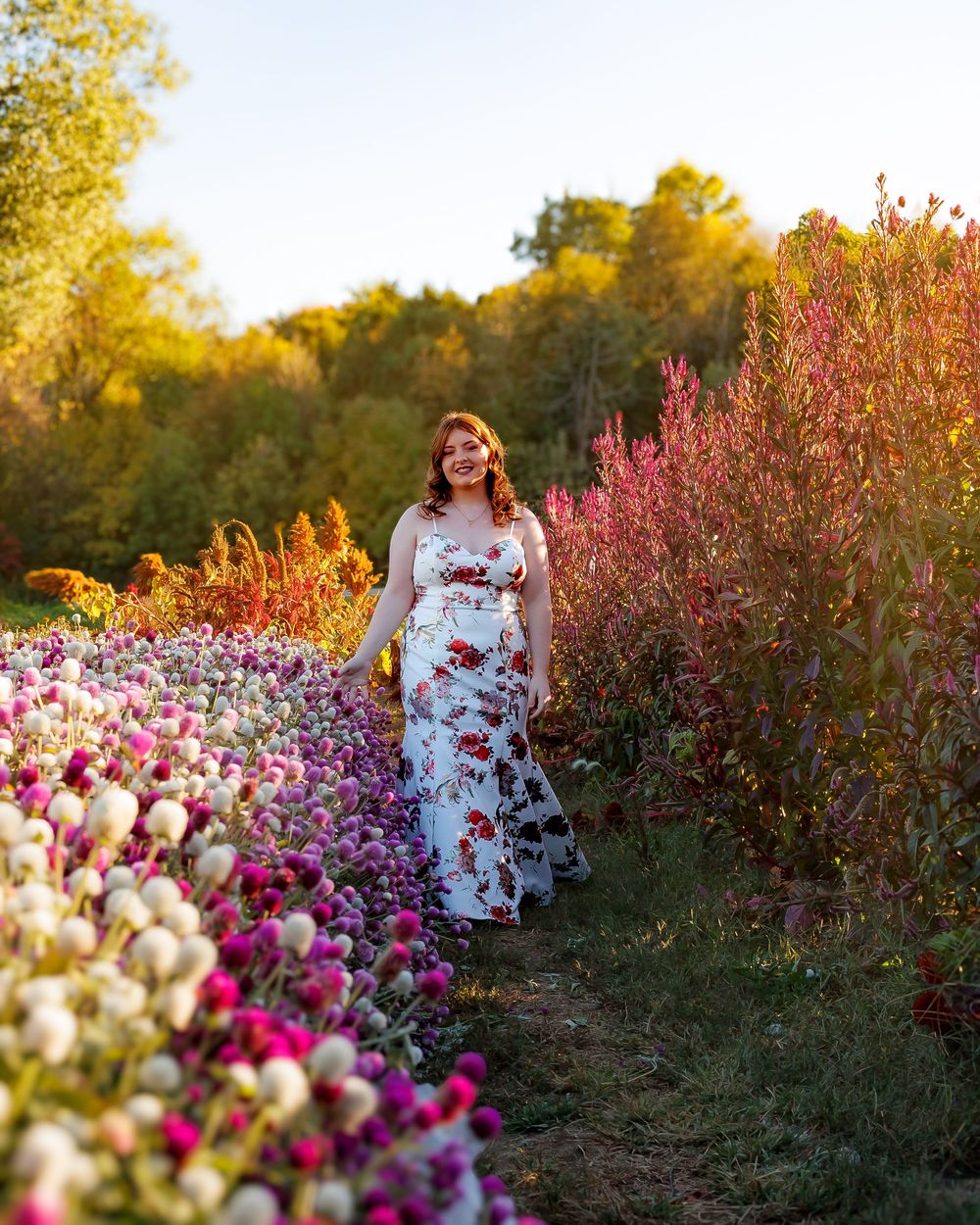 A person in a white floral dress walks through a vibrant garden path lined with pink and yellow blooms at sunset.