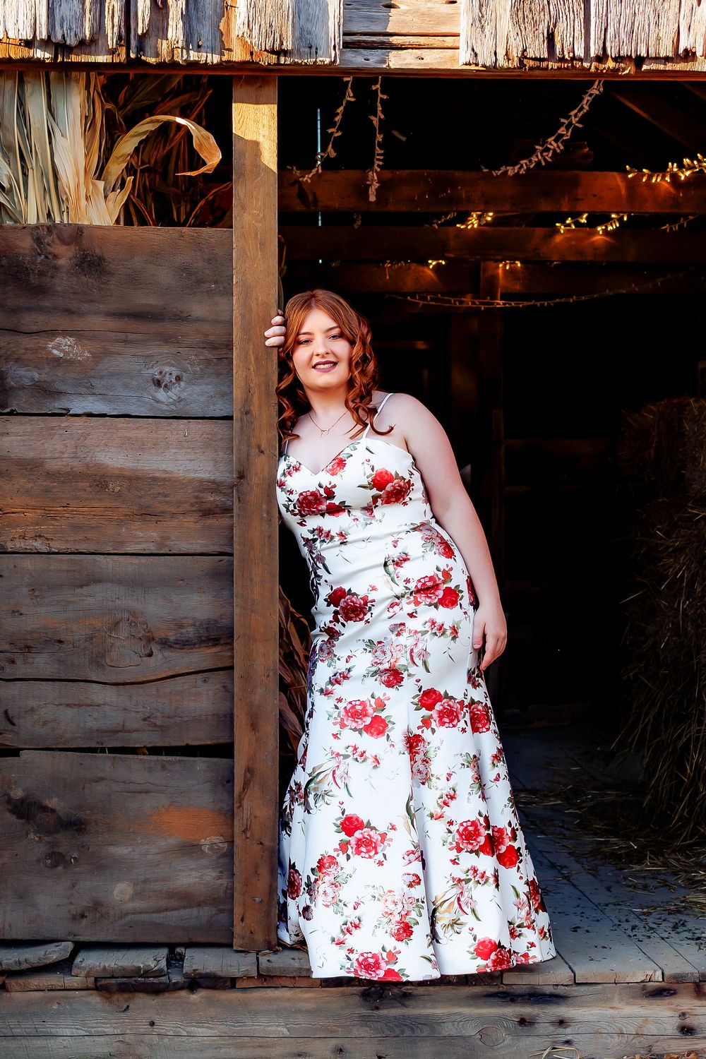 A floral white dress is showcased against a rustic wooden barn backdrop with warm lighting and string lights visible.