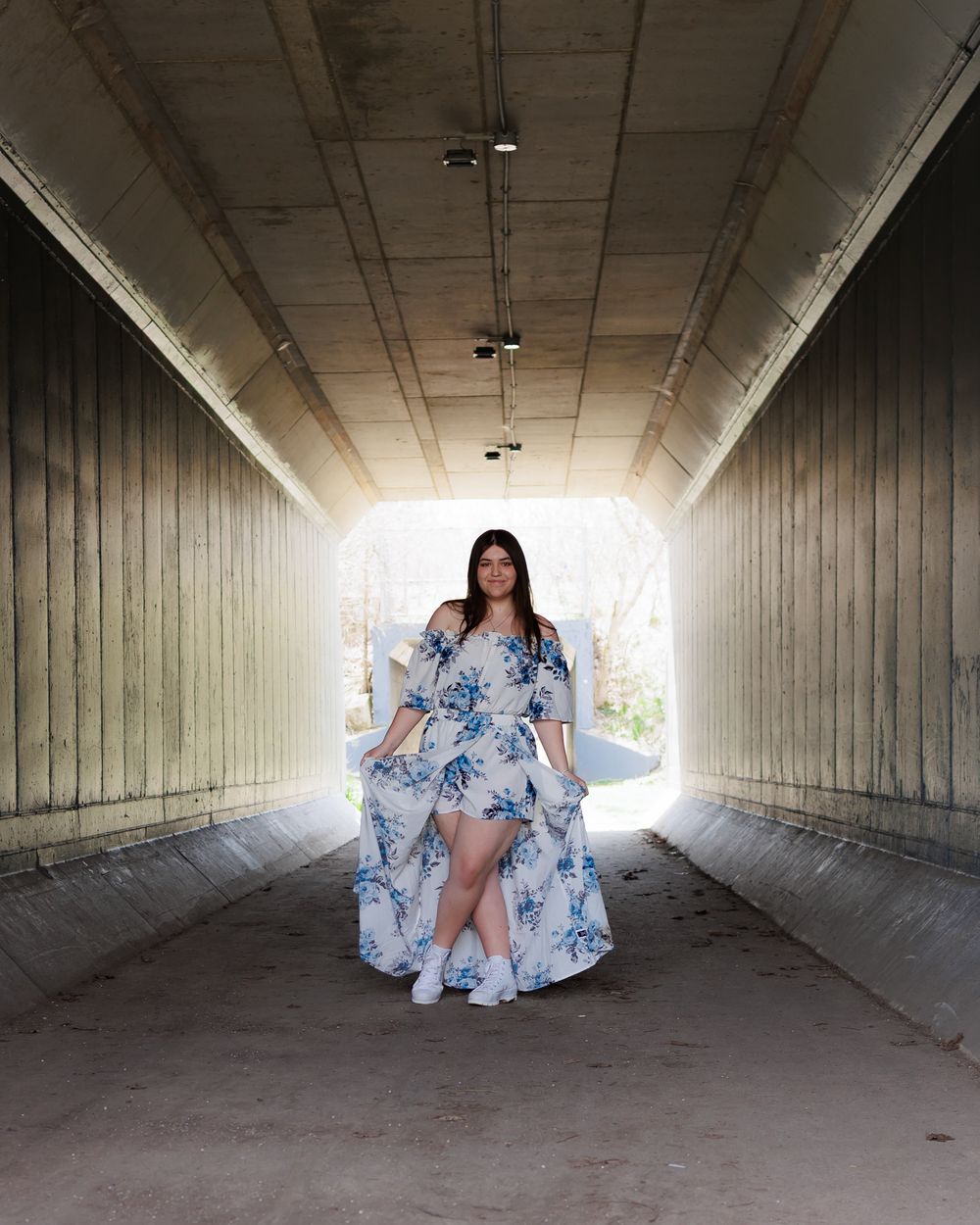 Someone in a flowing blue and white floral dress stands in a concrete tunnel with bright light at the end.