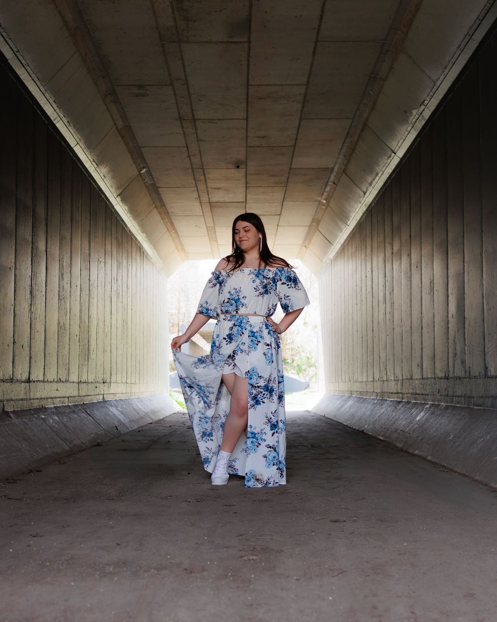 Person in a floral blue and white maxi dress poses in a concrete tunnel with dramatic backlit lighting.