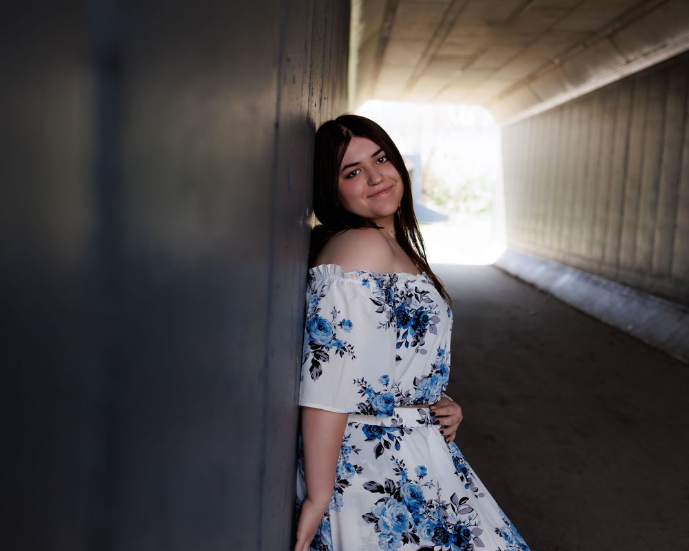 Someone in a white and blue floral off-shoulder dress poses in a tunnel with dramatic backlighting.