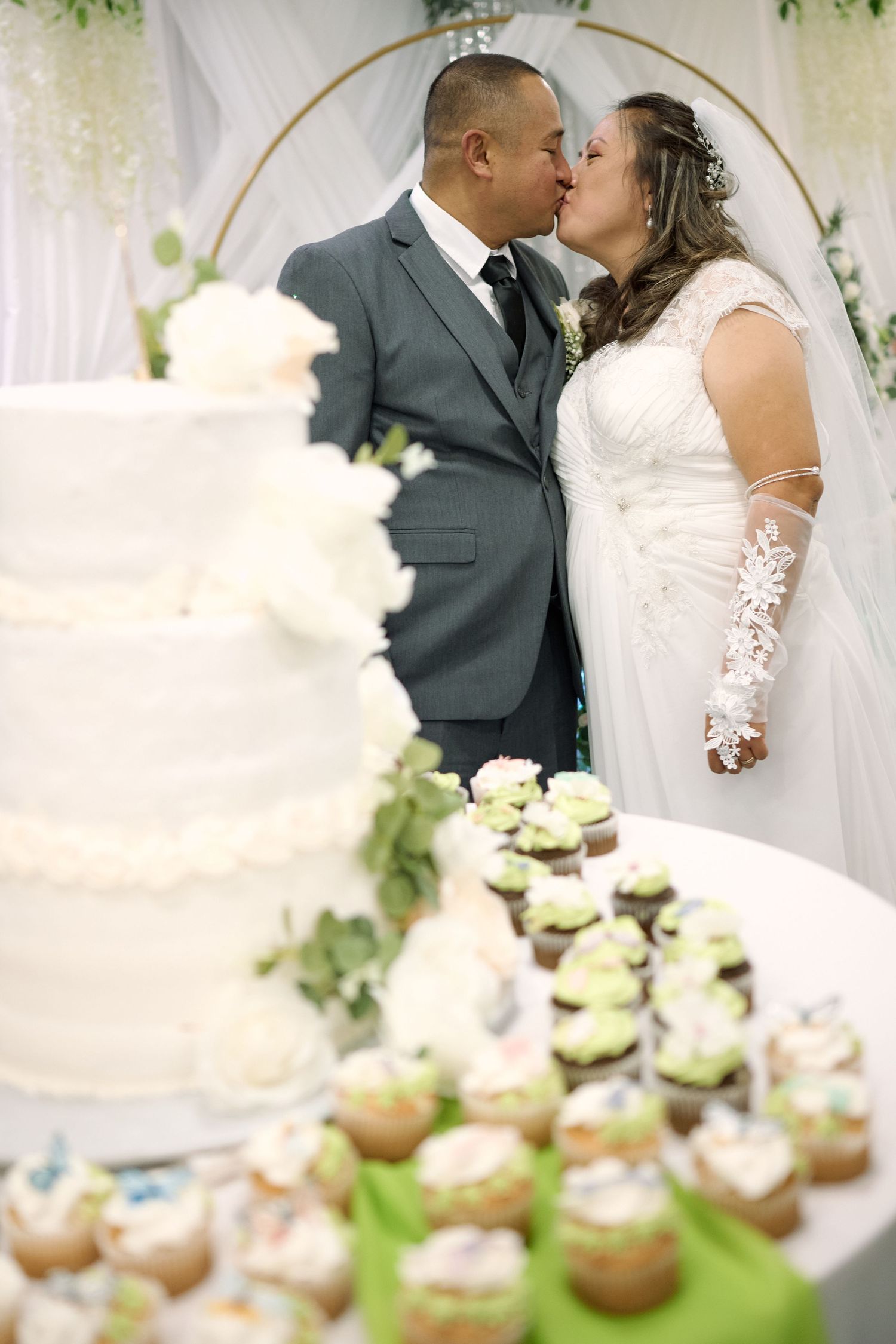 A wedding celebration with a beautiful white cake and decorated cupcakes displayed on a dessert table at the reception.