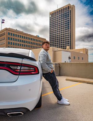 Person leaning on white car in urban parking lot with tall office building in background.