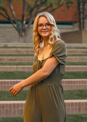 A person in a green peasant dress and glasses poses outdoors against stone steps during golden hour lighting.