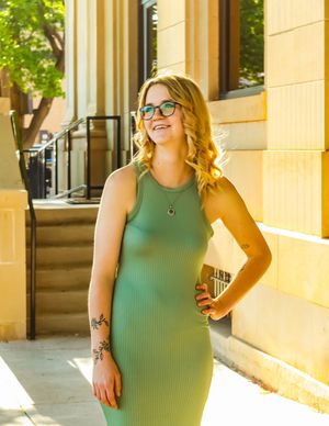 A person in a fitted green ribbed dress poses in front of a yellow building on a sunny day.