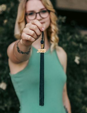 A hand holds up a black graduation tassel with a gold 2023 charm against a green outdoor background.