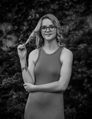 Black and white portrait of someone in glasses and a sleeveless ribbed top against a leafy background.