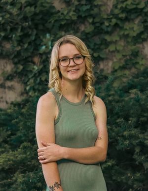 Portrait of someone in a ribbed sage green tank top and glasses standing in front of green foliage outdoors.